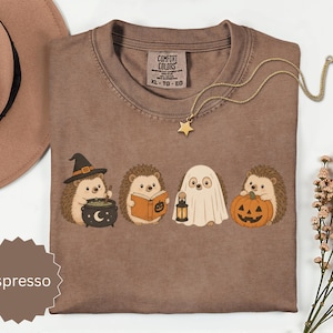 May include: An espresso-colored t-shirt featuring a graphic of four hedgehogs in Halloween costumes: one with a witch hat and cauldron, one reading a book, one dressed as a ghost holding a lantern, and one holding a jack-o'-lantern pumpkin.