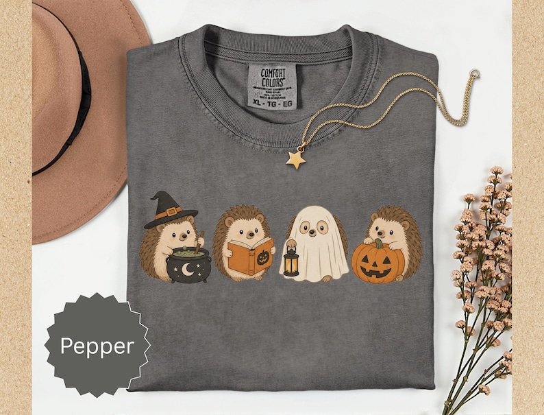 May include: A charcoal gray t-shirt featuring a graphic of four hedgehogs in Halloween costumes. One wears a witch hat with a cauldron, one reads a book, one is dressed as a ghost holding a lantern, and one holds a jack-o'-lantern pumpkin.
