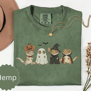 Peut inclure: Un t-shirt vert sauge avec un motif représentant quatre geckos déguisés pour Halloween : un avec une citrouille sur la tête, un en fantôme, un en sorcière avec un balai et un en vampire. Le t-shirt porte l'étiquette Comfort Colors et le mot Hemp.
