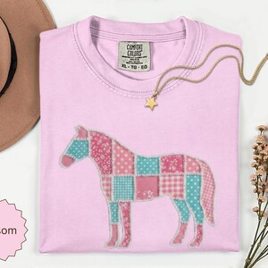 May include: A light pink t-shirt featuring a patchwork horse design in shades of pink and teal. The horse graphic is composed of various patterned squares. A gold star pendant necklace rests on the neckline, and a Comfort Colors label is visible.