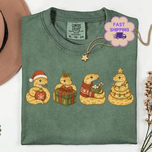 May include: A moss green t-shirt featuring a graphic of four yellow snakes. The first wears a Santa hat and holds an ornament, the second sits by a gift, the third wears a sweater and holds a mug, and the fourth is wrapped in Christmas lights.