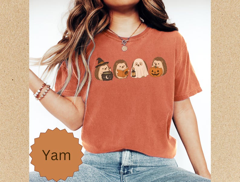May include: A rust-colored short-sleeve t-shirt featuring a graphic print of four hedgehogs in Halloween costumes, including a witch with a cauldron, a reader with a book, a ghost with a lantern, and one holding a pumpkin. The word Yam is printed below.