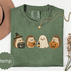 May include: A sage green t-shirt featuring a graphic of four hedgehogs in Halloween costumes: one with a witch hat and cauldron, one reading a book, one dressed as a ghost holding a lantern, and one holding a jack-o'-lantern pumpkin.