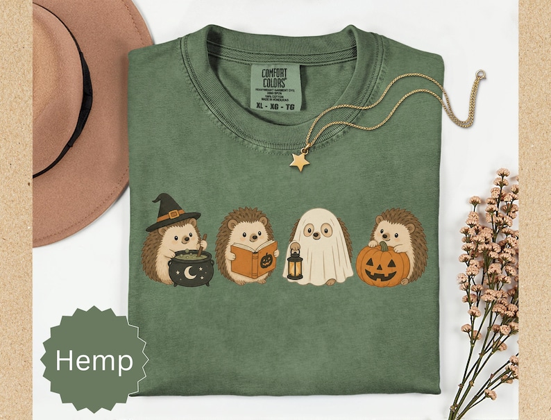 May include: A sage green t-shirt featuring a graphic of four hedgehogs in Halloween costumes: one with a witch hat and cauldron, one reading a book, one dressed as a ghost holding a lantern, and one holding a jack-o'-lantern pumpkin.