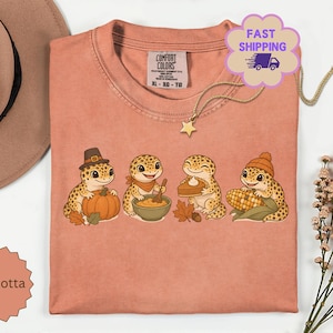 Könnte beinhalten: Ein terrakottafarbenes T-Shirt mit einer Grafik von vier Leopardgeckos. Einer trägt einen Pilgerhut mit einem Kürbis, einer trägt einen Schal und rührt in einer Schüssel, einer hält ein Stück Kuchen und einer trägt eine Mütze mit einem Maiskolben.