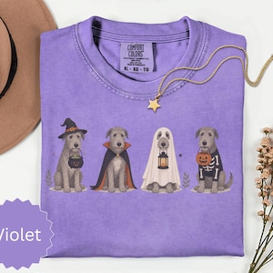 May include: A violet t-shirt featuring a graphic of four dogs in Halloween costumes: a witch with a cauldron, a vampire in a cape, a ghost with a lantern, and a skeleton with a pumpkin bucket. The shirt has a Comfort Colors label in the neck.
