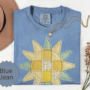 May include: A blue jean coloured t-shirt featuring a central patchwork sunflower design made of yellow and green fabric squares with frayed edges. The shirt has a Comfort Colors label and is accompanied by a gold star pendant necklace.