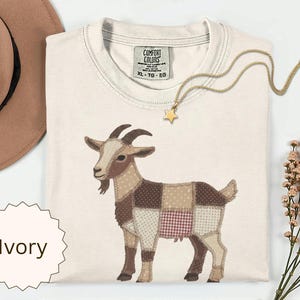 May include: An ivory t-shirt featuring a graphic of a goat designed with a patchwork pattern of brown, beige, and red checkered fabrics. The shirt has a Comfort Colors label at the neck with size XL, TG, EG printed. A gold star pendant necklace is included.