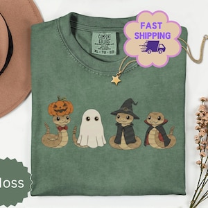 Może przedstawiać: Mchowo-zielony t-shirt z grafiką przedstawiającą cztery węże przebrane na Halloween: jeden z głową dyni, jeden jako duch, jeden w kapeluszu czarownicy i pelerynie oraz jeden w pelerynie wampira. Koszulka ma okrągły dekolt i metkę Comfort Colors.