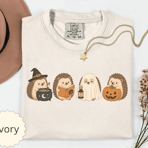 May include: An ivory t-shirt featuring a graphic of four hedgehogs in Halloween costumes: one with a witch hat and cauldron, one reading a book, one dressed as a ghost holding a lantern, and one holding a jack-o'-lantern pumpkin.