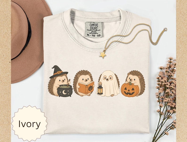 May include: An ivory t-shirt featuring a graphic of four hedgehogs in Halloween costumes: one with a witch hat and cauldron, one reading a book, one dressed as a ghost holding a lantern, and one holding a jack-o'-lantern pumpkin.