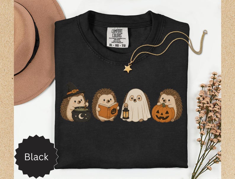 May include: A black t-shirt featuring a graphic of four hedgehogs in Halloween costumes. One wears a witch hat with a cauldron, one reads a book, one is dressed as a ghost holding a lantern, and one holds a jack-o'-lantern. The shirt has a Comfort Colors label.