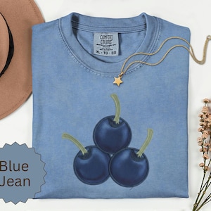 Op de afbeelding: Een blauw spijkerkleurig t-shirt met een geborduurde afbeelding van drie donkerblauwe kersen met lichtgroene steeltjes. Het shirt heeft een Comfort Colors-label in de nek, maat XL, en wordt getoond met een gouden ster-hanger.