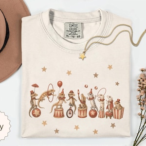 Puede incluir: Una camiseta de color marfil con un estampado gráfico de varios lagartos realizando actos de circo, incluyendo equilibrio sobre pelotas, montar en monociclo y usar sombreros de copa. La camiseta tiene una etiqueta de Comfort Colors en el cuello.