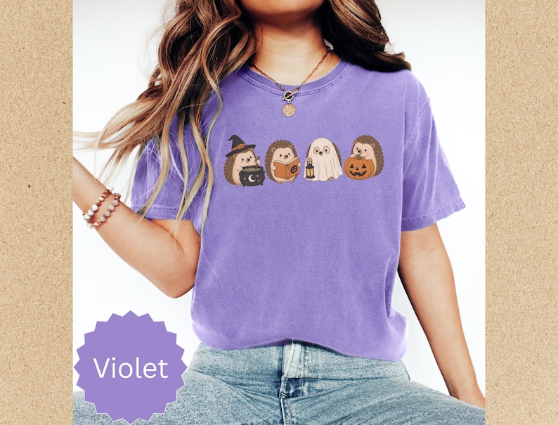 May include: A violet short-sleeve t-shirt featuring a graphic print of four hedgehogs in Halloween costumes. One hedgehog wears a witch hat with a cauldron, one reads a book, one is dressed as a ghost with a lantern, and one holds a pumpkin.