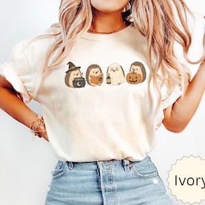 May include: An ivory short-sleeve t-shirt featuring a graphic print of four hedgehogs in Halloween costumes. The hedgehogs are depicted as a witch with a cauldron, a reader with a book, a ghost with a lantern, and one holding a jack-o'-lantern.