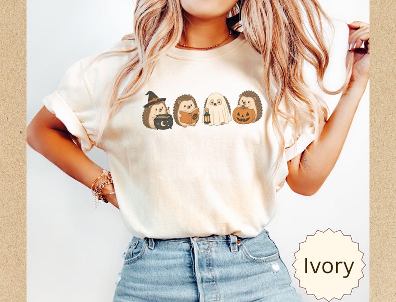 May include: An ivory short-sleeve t-shirt featuring a graphic print of four hedgehogs in Halloween costumes. The hedgehogs are depicted as a witch with a cauldron, a reader with a book, a ghost with a lantern, and one holding a jack-o'-lantern.