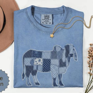 Peut inclure: Un t-shirt bleu jean avec un motif de vache composé de divers patchs en tissu à motifs bleus et blancs. Le t-shirt porte une étiquette Comfort Colors au col et un collier avec un pendentif étoile dorée est posé sur le tissu.