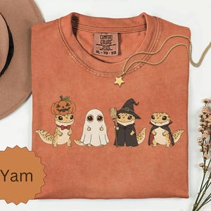 May include: A rust-coloured t-shirt featuring a graphic of four geckos in Halloween costumes: one with a pumpkin, one as a ghost, one as a witch with a broom, and one as a vampire. The shirt has a Comfort Colors label with size XL-TG-EG text.