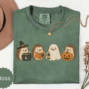 May include: A moss green t-shirt featuring a graphic of four hedgehogs in Halloween costumes: one with a witch hat and cauldron, one reading a book, one dressed as a ghost holding a lantern, and one holding a jack-o'-lantern pumpkin.