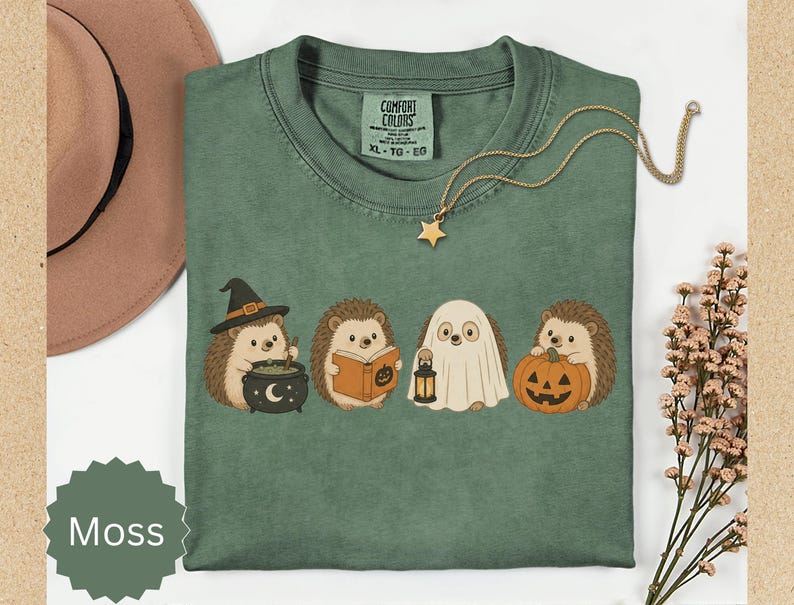 May include: A moss green t-shirt featuring a graphic of four hedgehogs in Halloween costumes: one with a witch hat and cauldron, one reading a book, one dressed as a ghost holding a lantern, and one holding a jack-o'-lantern pumpkin.