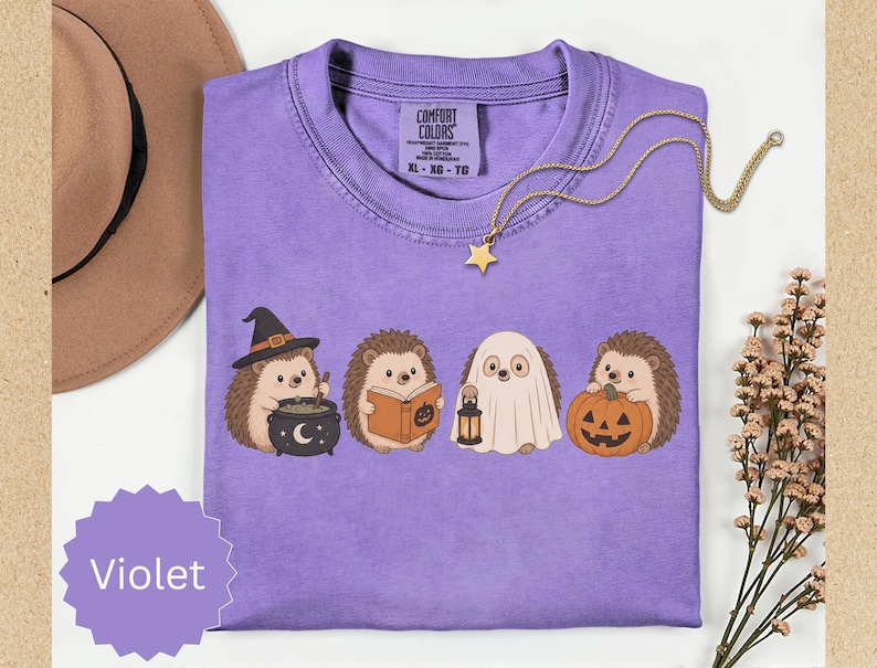 May include: A violet short-sleeved t-shirt featuring a graphic of four hedgehogs in Halloween costumes. One wears a witch hat with a cauldron, one reads a book, one is dressed as a ghost holding a lantern, and one holds a jack-o'-lantern pumpkin.