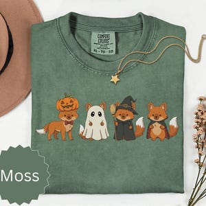 Peut inclure: Un t-shirt à col rond vert mousse avec un motif représentant quatre renards en costumes d'Halloween : un avec une citrouille sur la tête, un en fantôme, un en sorcière avec un balai et un en vampire. Le t-shirt porte une étiquette Comfort Colors.