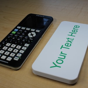 May include: A black and silver graphing calculator with a white case that says "Your Text Here" in green letters.
