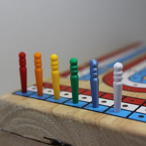 May include: A wooden cribbage board with colorful pegs. The pegs are red, orange, yellow, green, blue, and white. The board has a red, white, and blue track design. The board is made of wood and has a natural finish.