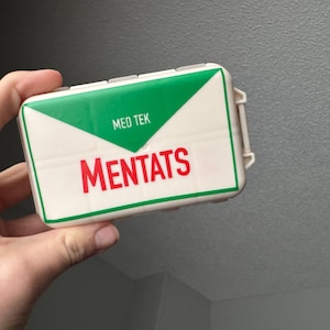 May include: A white rectangular box with a green triangle design on the top. The box has the text "MED TEK" and "MENTATS" in red letters. The box is held in a hand.