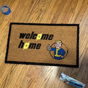 May include: A rectangular doormat with the words "welcome home" in black, with yellow lightning bolt accents. A cartoon character in a blue and yellow jumpsuit gives a thumbs up. The mat has a black border.