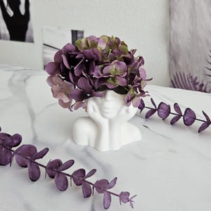 May include: A white ceramic planter shaped like a woman's face with purple flowers in her hair. The planter is sitting on a white surface.