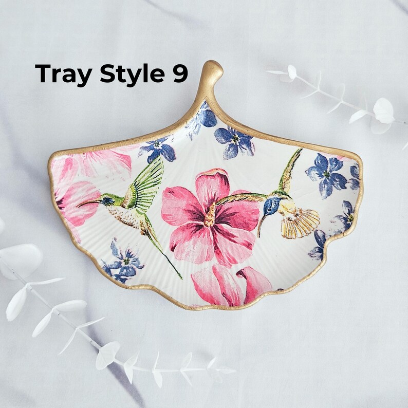 May include: A decorative, fan-shaped ceramic tray with a gold rim. The tray features a floral design with pink hibiscus flowers, blue flowers, and hummingbirds. The text "Tray Style 9" is printed on the top left.