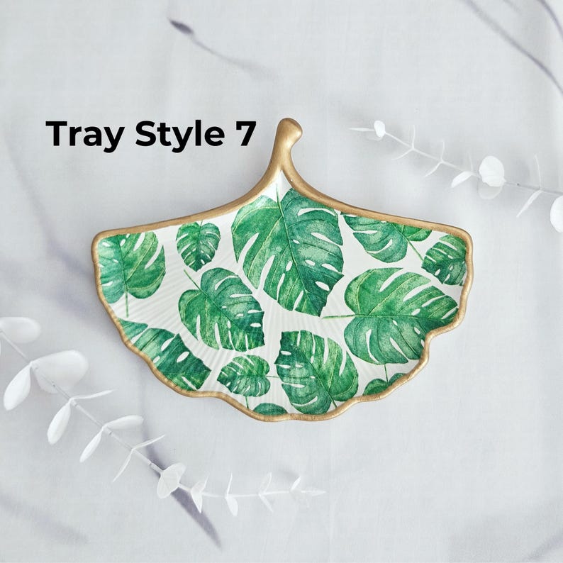 May include: A decorative, leaf-shaped tray with a gold trim and a white background. The tray is adorned with a pattern of green monstera leaves. The text "Tray Style 7" is visible on the left side of the tray.