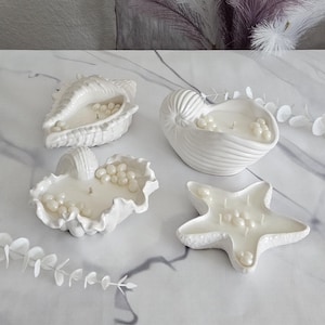 May include: Four white seashell-shaped candles with pearl-like accents. The candles are in the shape of a conch shell, nautilus shell, scallop shell, and starfish. The candles are on a white marble surface.
