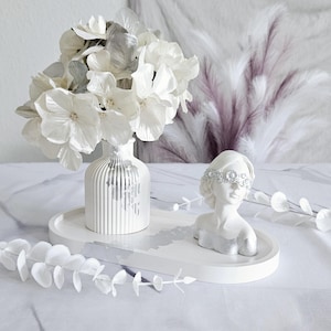 May include: White ceramic tray with a white vase filled with white and silver flowers and a white ceramic bust with a silver headband. The tray is on a white surface.