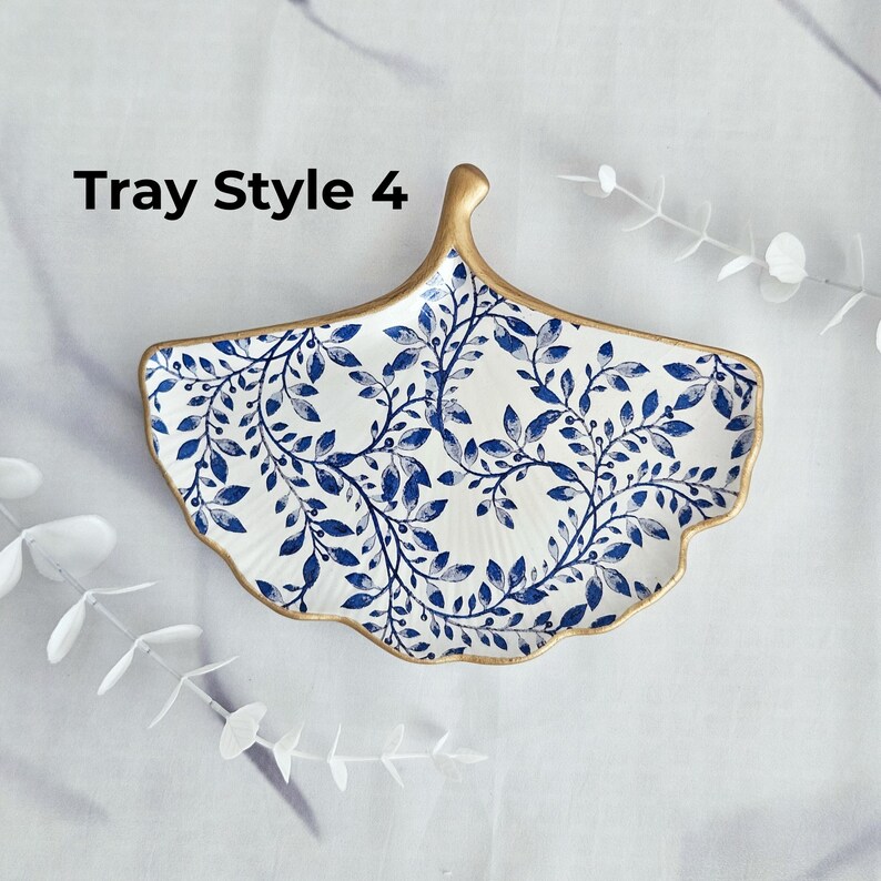 May include: A decorative ceramic tray in a fan shape, featuring a white background with a blue leaf pattern. The rim and handle are trimmed with gold. The text "Tray Style 4" is visible.