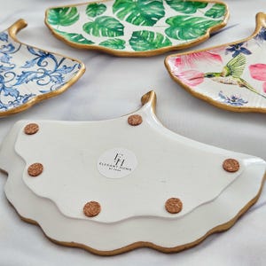 May include: A collection of decorative ceramic trays with gold trim. Designs include a blue floral pattern, green monstera leaves, pink flowers with a hummingbird, and a white tray with the text "ELEGANT HOME BY TARA".