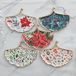 May include: Five decorative, leaf-shaped ceramic plates with gold trim. Each plate features a different floral or botanical design, including poinsettias, berries, and various flowers. The plates are arranged on a white marble surface.
