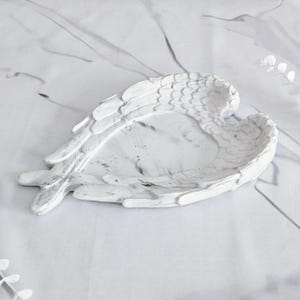 May include: White and gray decorative angel wing tray. The tray is shaped like a pair of wings with detailed feathering. The interior of the tray has a marbled design. The tray is made of a solid material and is suitable for holding small items.