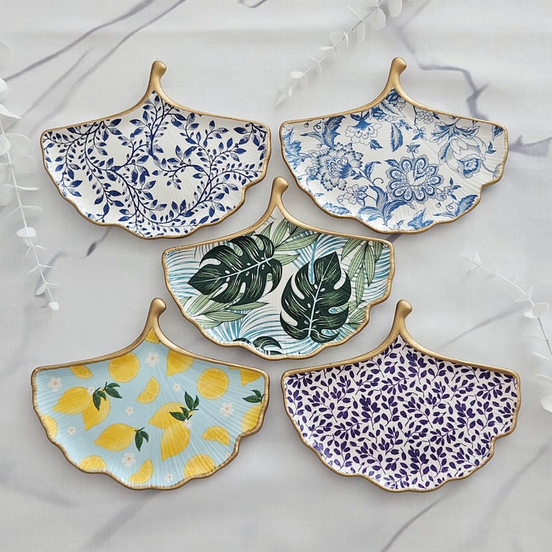 May include: Five decorative leaf-shaped ceramic dishes with gold trim. The dishes feature various patterns, including blue floral, green tropical leaves, yellow lemons, and blue and white foliage. The dishes are arranged on a white marble surface.