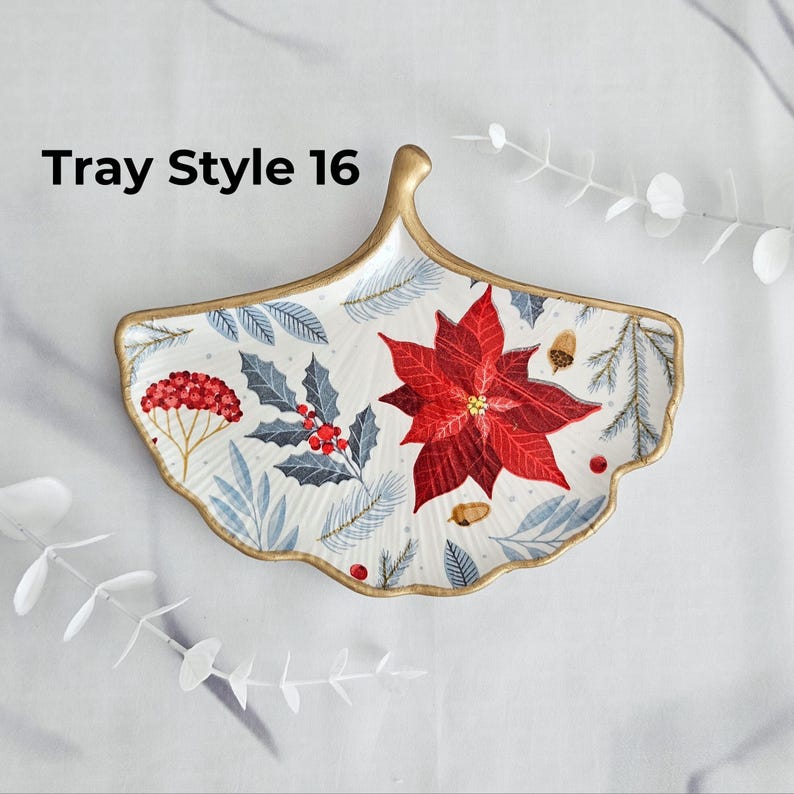 May include: A decorative, leaf-shaped tray with a gold rim and handle. The tray features a poinsettia, holly, and pine needle design in red, blue, and white. The text "Tray Style 16" is visible on the upper left.