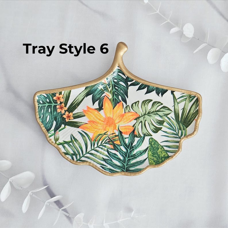 May include: A decorative, leaf-shaped tray with a gold rim and a tropical floral design. The tray features a large orange flower and green leaves on a white background. The text "Tray Style 6" is visible.