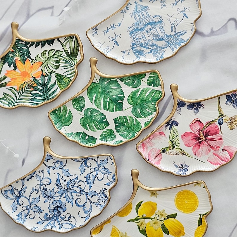 May include: A collection of decorative, fan-shaped plates with gold trim. The plates feature various botanical and scenic designs, including tropical leaves, flowers, and a blue and white pagoda scene. The plates are white with colorful designs.