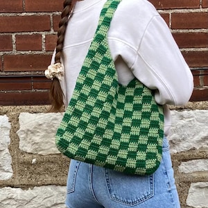 May include: A green and light green checkered crochet tote bag with a single strap. The bag is worn over the shoulder of a person wearing blue jeans and a white sweater.