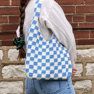 Peut inclure: Un sac cabas en crochet à carreaux bleus et blancs avec une seule sangle. Le sac est porté sur l'épaule d'une personne portant un jean bleu et un pull blanc.