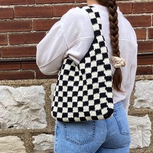 May include: A black and white checkered crochet tote bag with a single strap. The bag is worn over the shoulder of a person wearing a white sweater and blue jeans.
