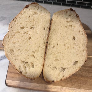 May include: Two halves of a loaf of bread, freshly sliced, revealing a light-colored interior with numerous air pockets. The crust is a golden brown. The bread rests on a wooden cutting board.