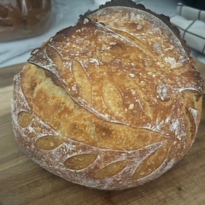 May include: A golden-brown loaf of sourdough bread with a leaf pattern cut into the crust. The bread sits on a wooden cutting board. Another loaf of bread is visible in the background.