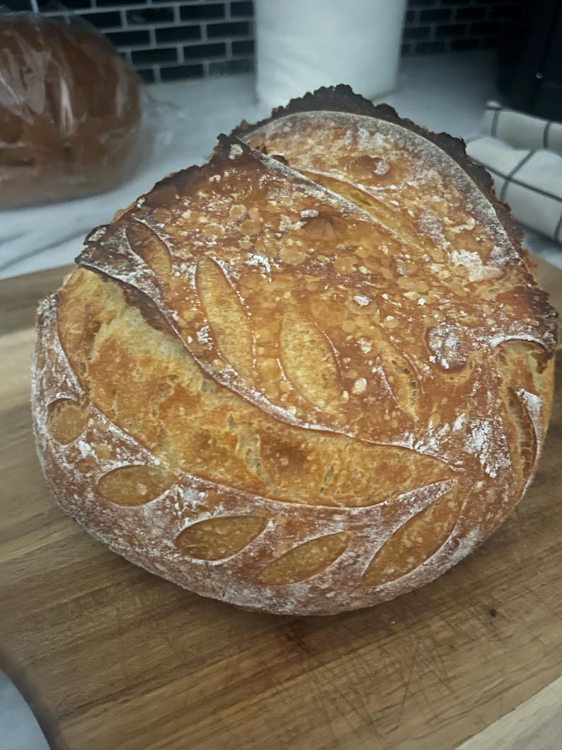 May include: A golden-brown loaf of sourdough bread with a leaf pattern cut into the crust. The bread sits on a wooden cutting board. Another loaf of bread is visible in the background.